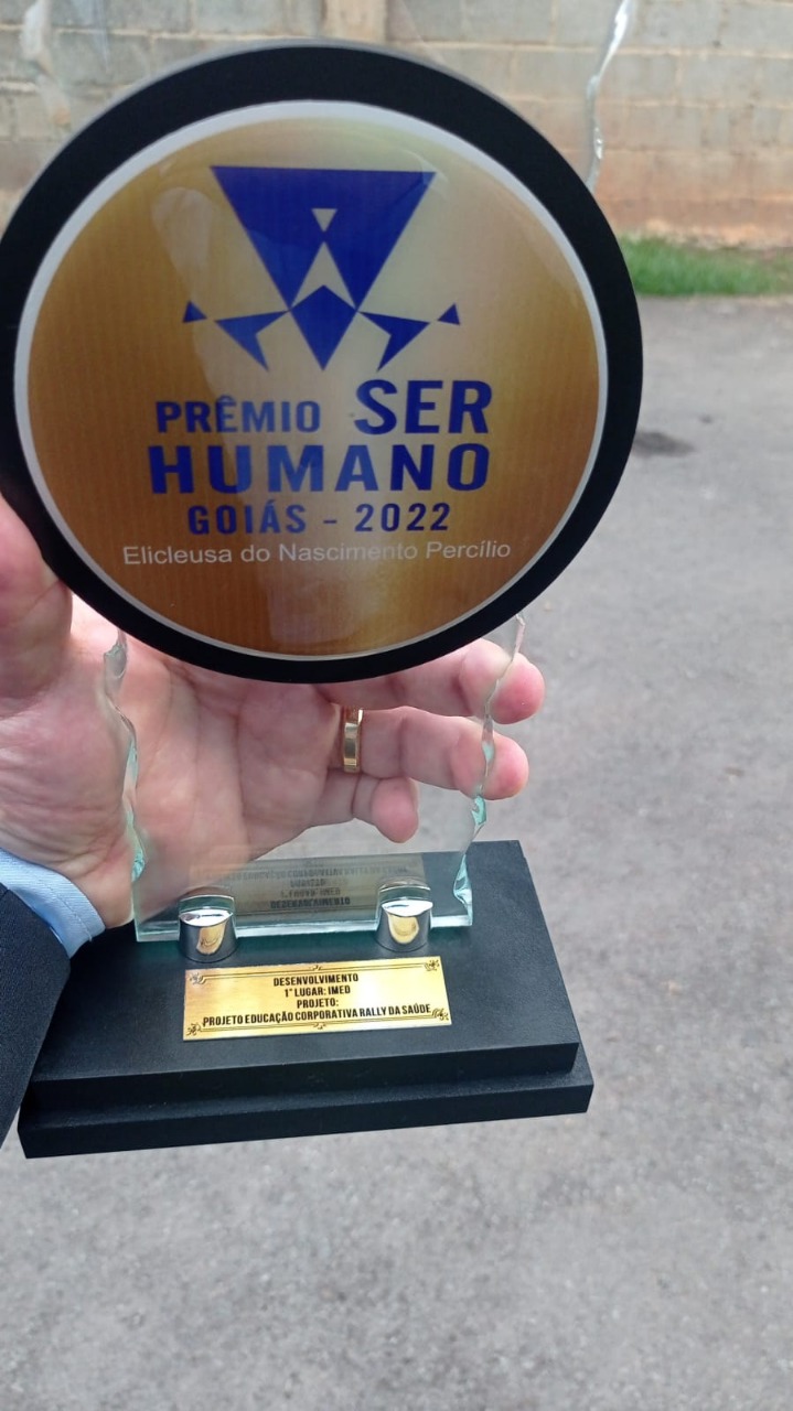 Registro de 4 pessoas segurando o prêmio Ser Humano. Um coqueiro ao lado esquerdo e uma tela azul com qr code ao fundo.
